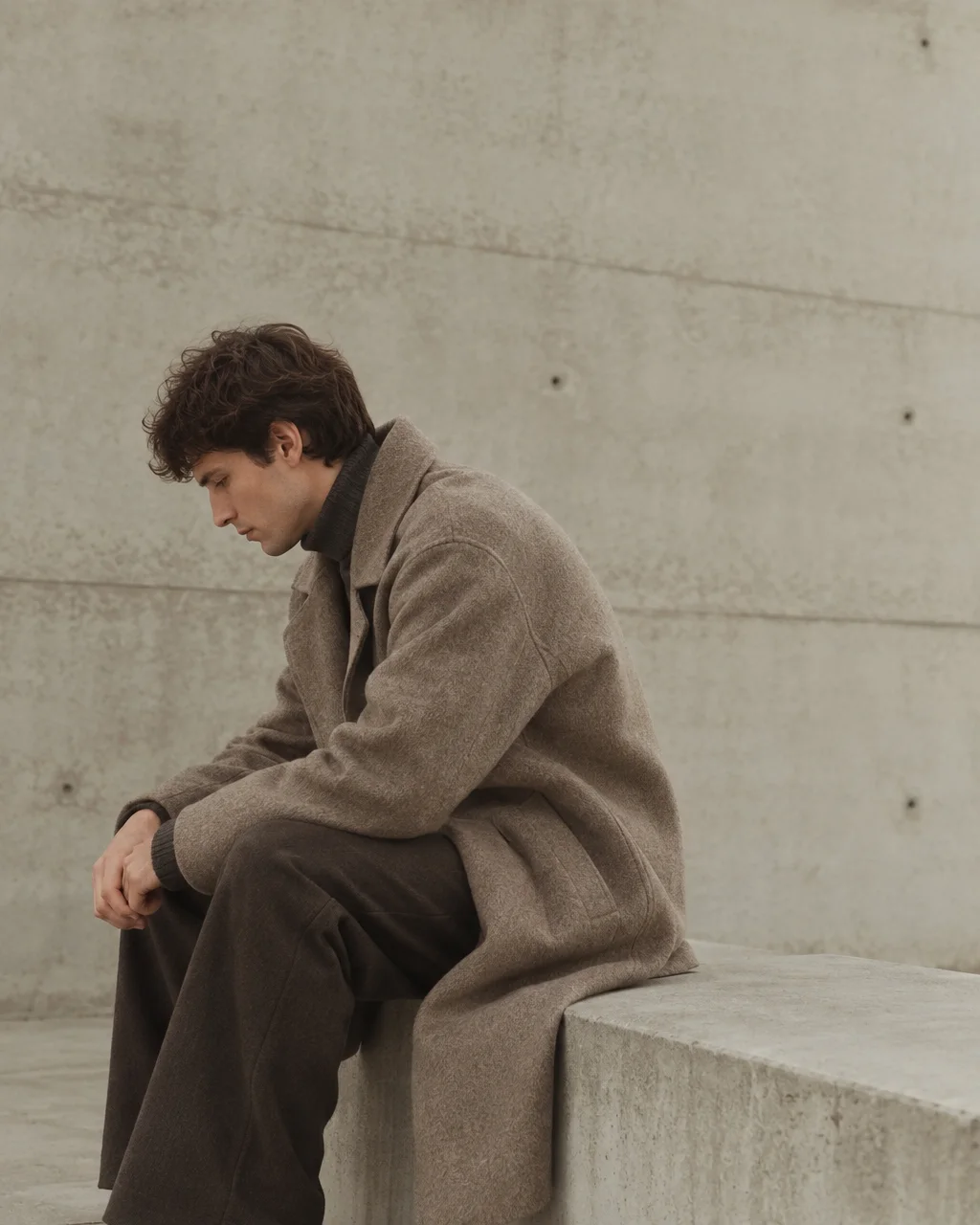 A man dressed in a warm wool overcoat sits quietly in contemplation against a minimalist architectural concrete wall.