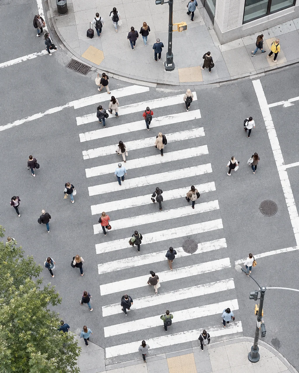 A high-angle view of pedestrians crossing a wide urban intersection with bold white painted lines across grey asphalt.