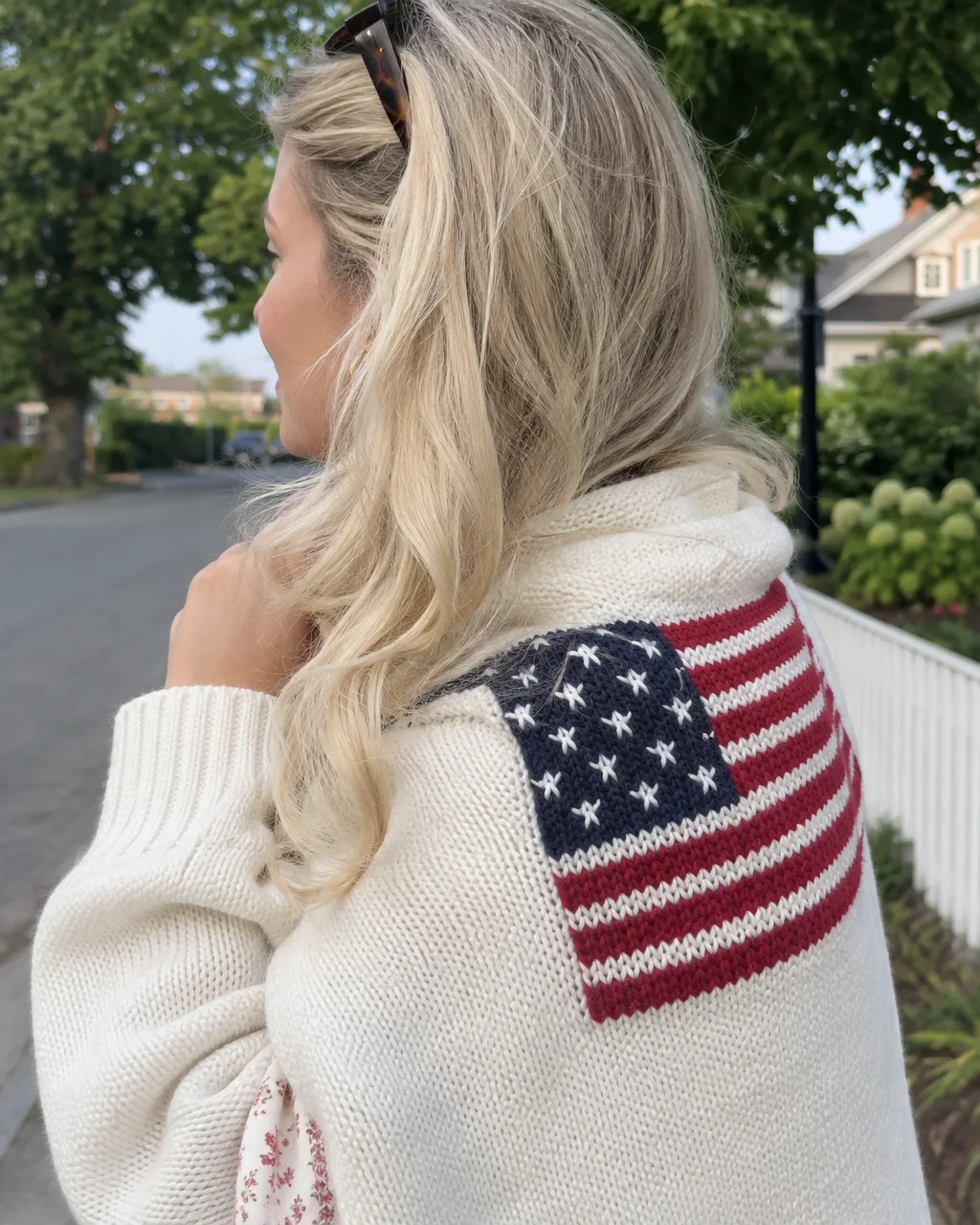 A blonde woman stands outdoors wearing a cream knit sweater featuring an American flag motif stitched across the shoulder.