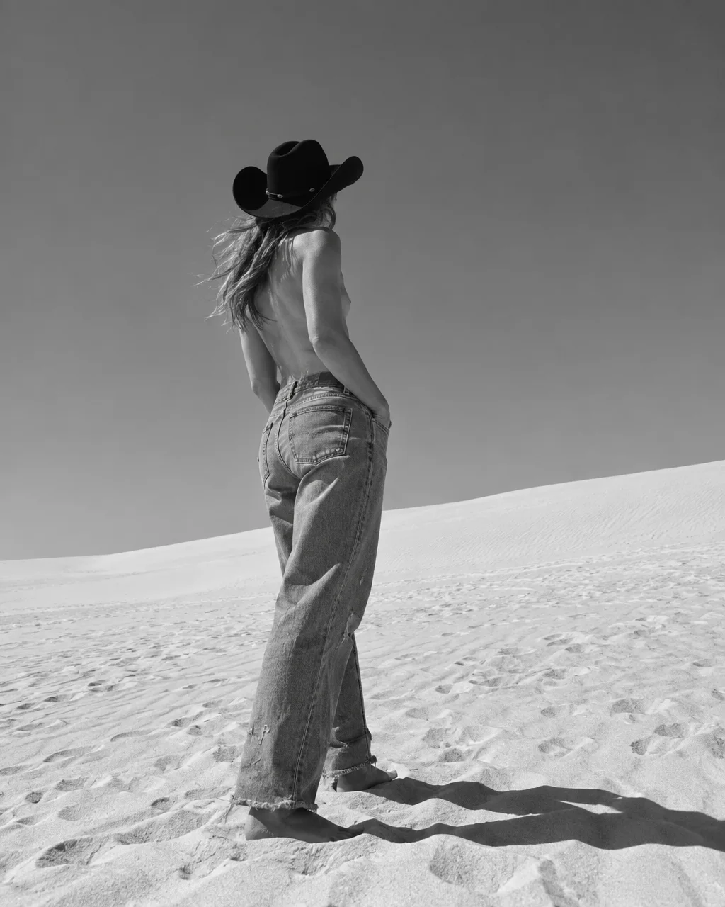 A black and white photograph of a woman wearing a black cowboy hat and jeans, standing barefoot across bright desert sand dunes.